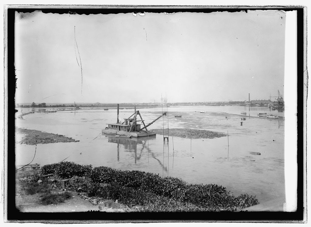 A steam dipper dredge at work among the mud flats of the Anacostia River in 1912, the Washington shoreline beyond.